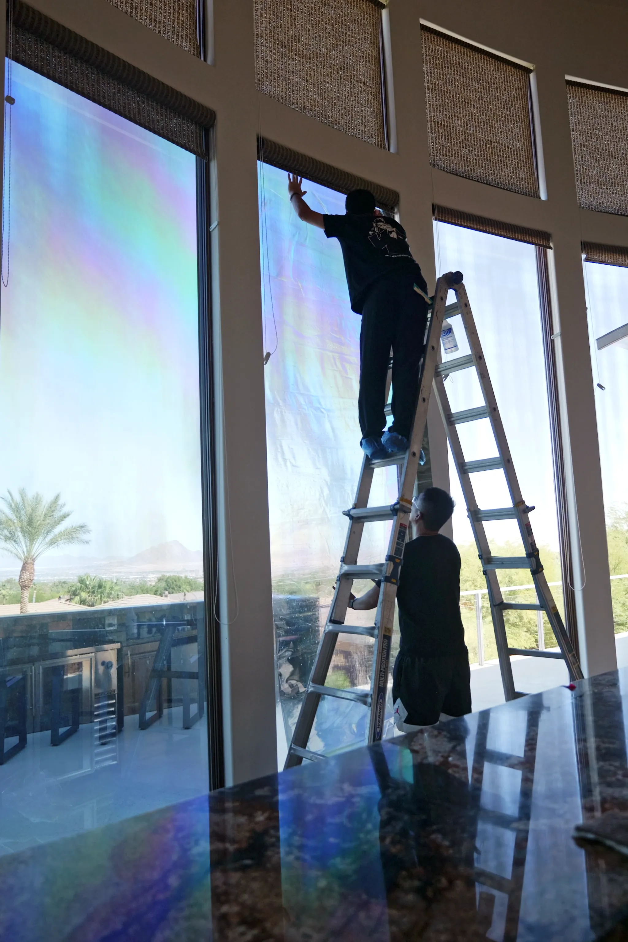 AT Customs installer on a ladder applying film to a tall window, the film still showing its rainbow sheen